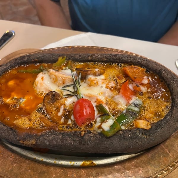 A bubbling Turkish casserole with melted cheese, tomatoes, and vegetables in a dark stone dish.