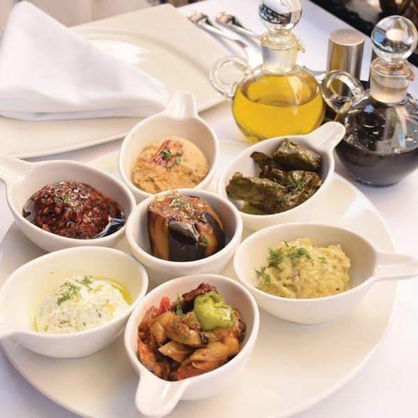 A white platter holds seven small bowls of various Turkish mezes, with oil and vinegar cruets nearby.